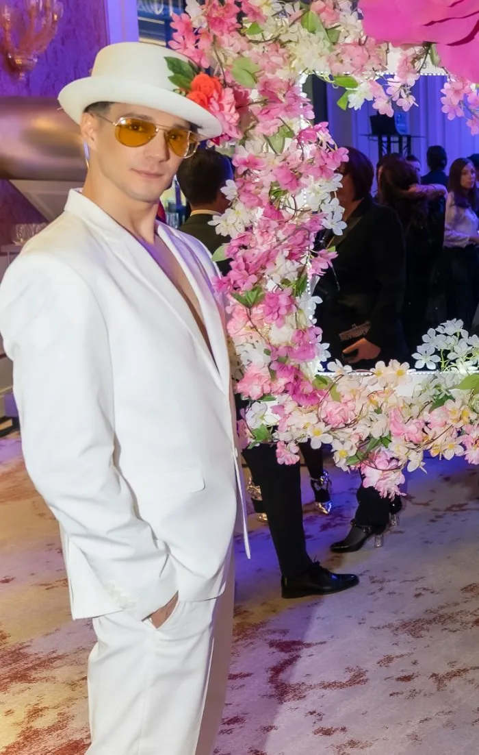 Artista con traje blanco y sombrero junto a un marco de flores en la bienvenida de un evento de empresa