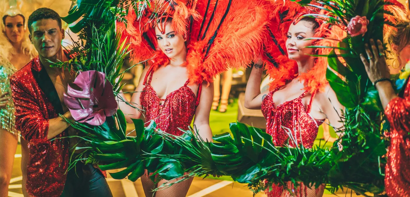 Bailarinas con plumas naranjas y un artista con chaqueta de lentejuelas tras un marco de hojas tropicales