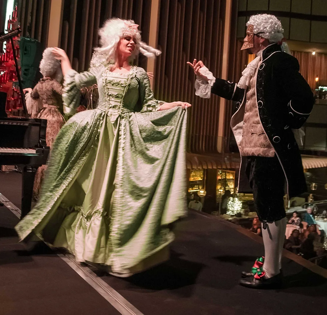 Actriz con vestido de corte verde y actor con casaca y máscara bailando en una cena temática de empresa