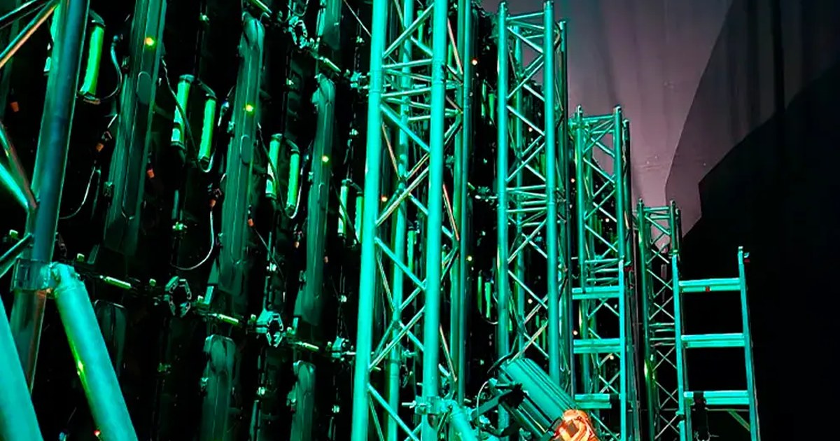 Parte trasera de una pantalla LED de gran formato sostenida por torres de truss, con un foco y una escalera de acceso