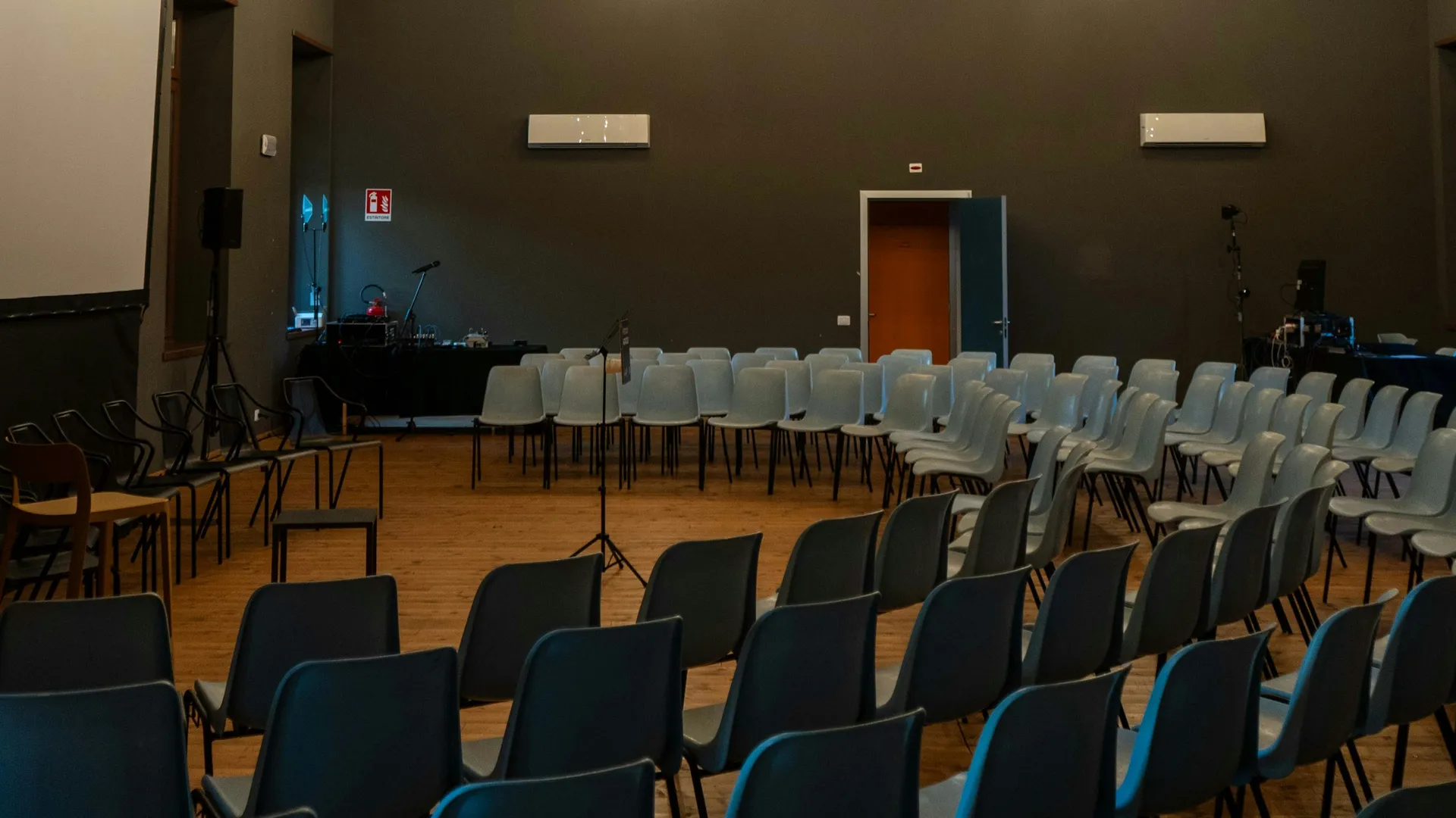 Sala con techo de vigas de madera y sillas en filas frente a una pantalla de proyección, con altavoz, micrófonos y mesa técnica preparados antes de una sesión