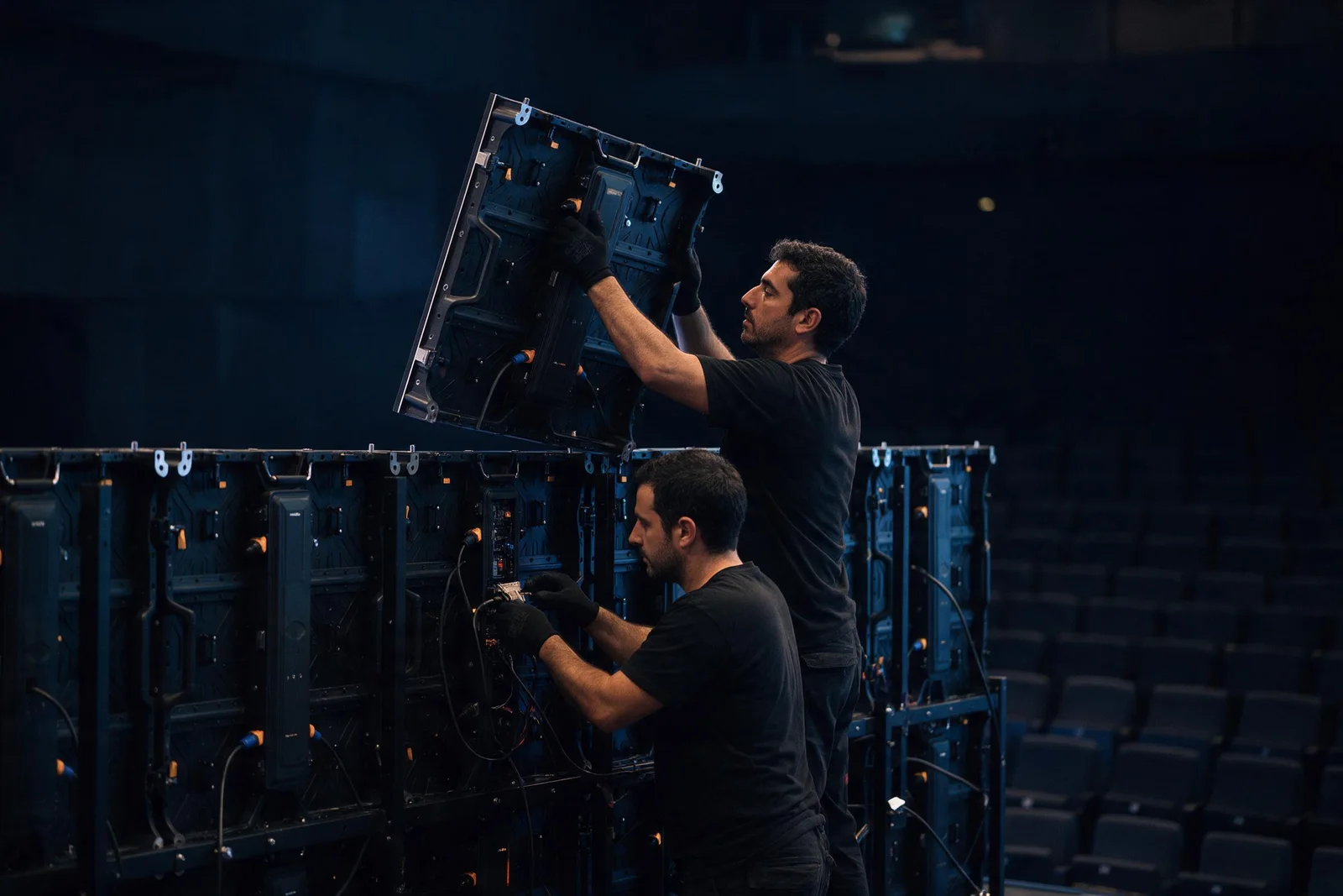 Dos técnicos montando los módulos de una pantalla LED sobre su estructura en un auditorio vacío