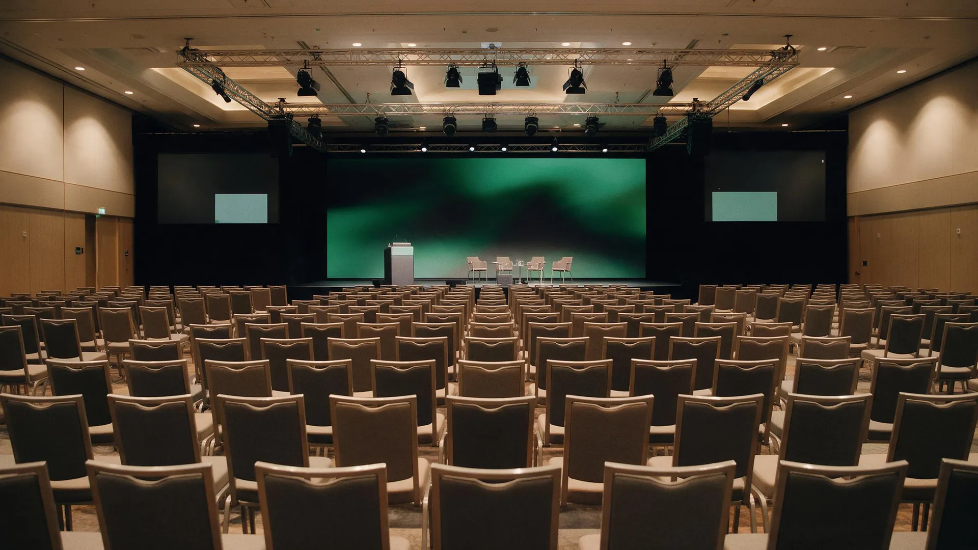 Sala plenaria vacía preparada para un congreso de empresa, con filas de sillas, atril y mesa de ponentes sobre el escenario, pantalla LED central, pantallas laterales y truss de iluminación