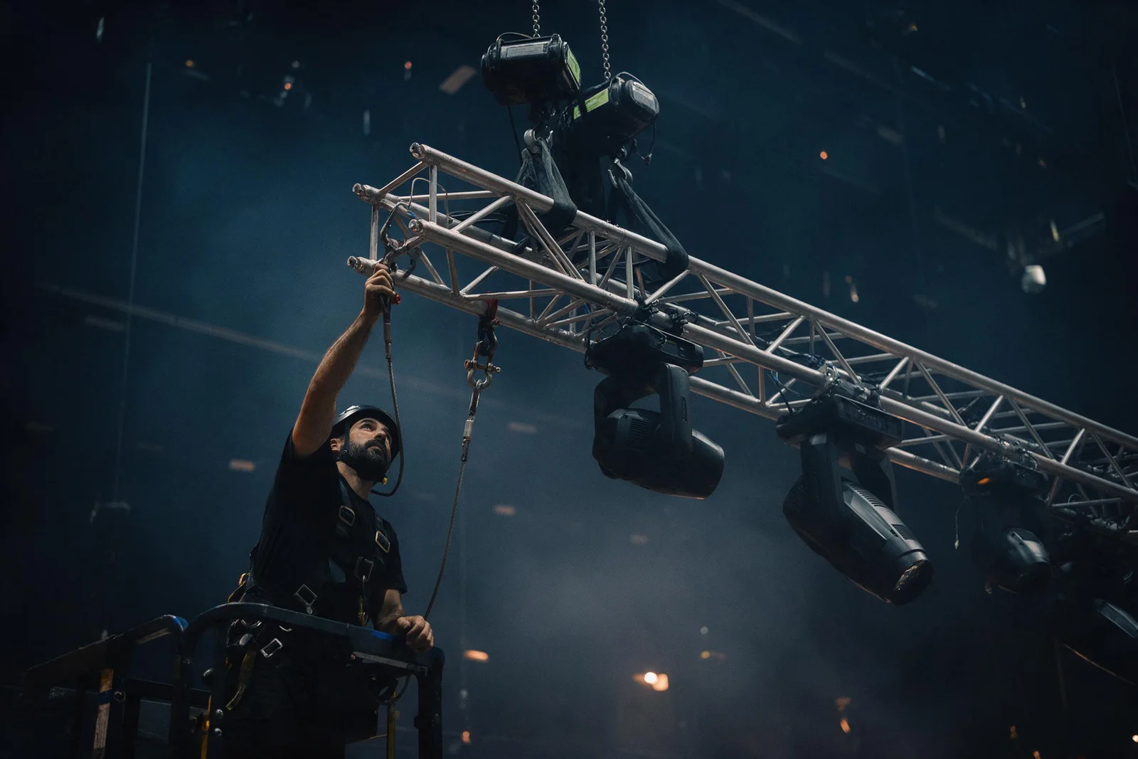 Técnico de rigging con arnés y casco revisando el colgado de un truss con cabezas móviles en un escenario