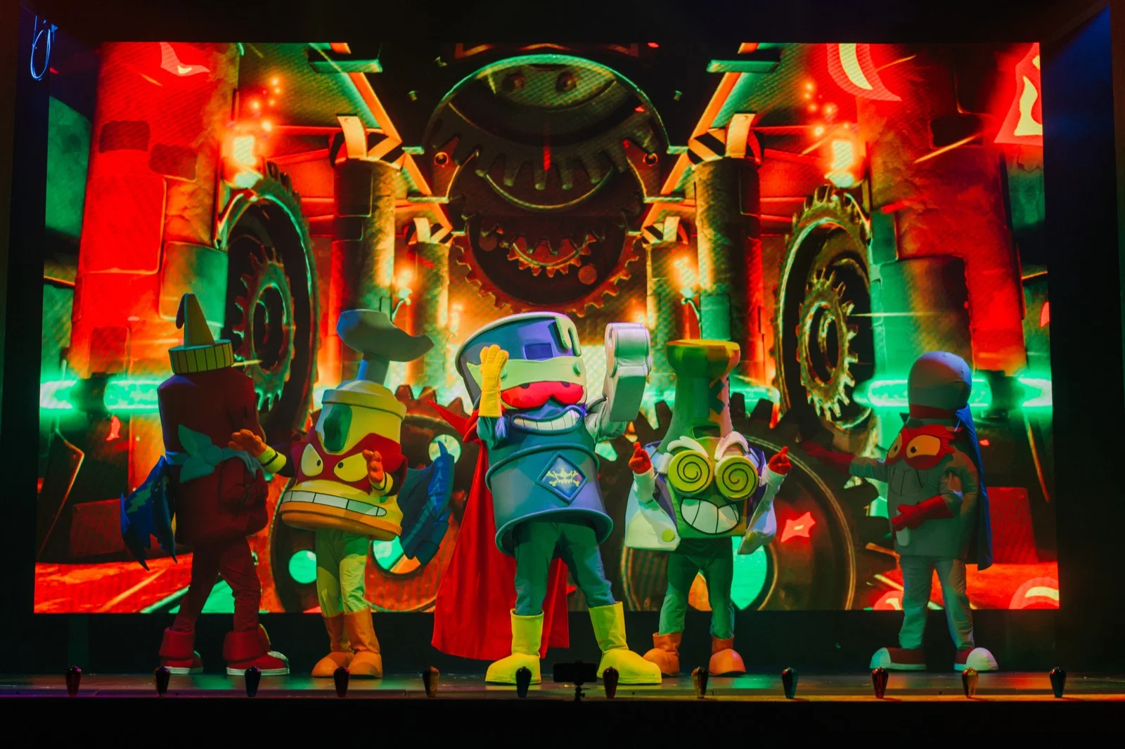Personajes de SuperThings en el escenario ante una pantalla LED con engranajes en tonos rojos y naranjas