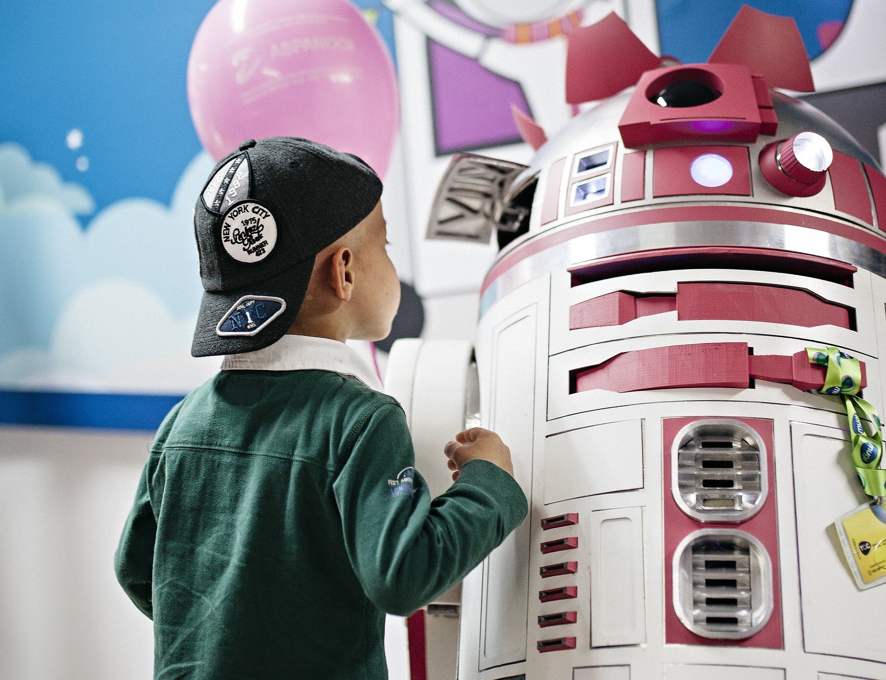 Un niño de espaldas, con gorra, observa de cerca al droide rosa R2-KT