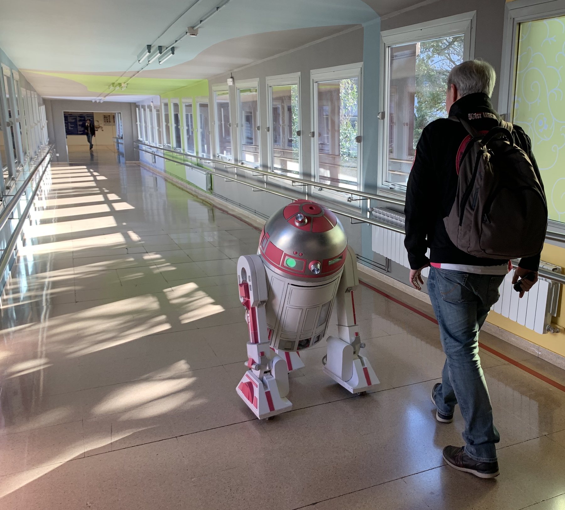 El droide rosa R2-KT recorre el pasillo luminoso de un hospital acompañado por un voluntario